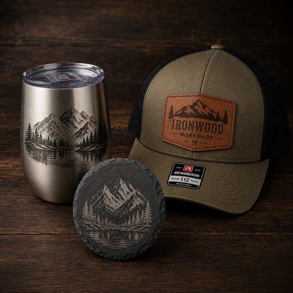 A mountain lake engraving design shown across three products: a brushed silver tumbler with a clear lid, a tan trucker cap with a brown leather patch reading Ironwood Workshop, and a dark slate coaster. The items are arranged on a dark wood surface.
