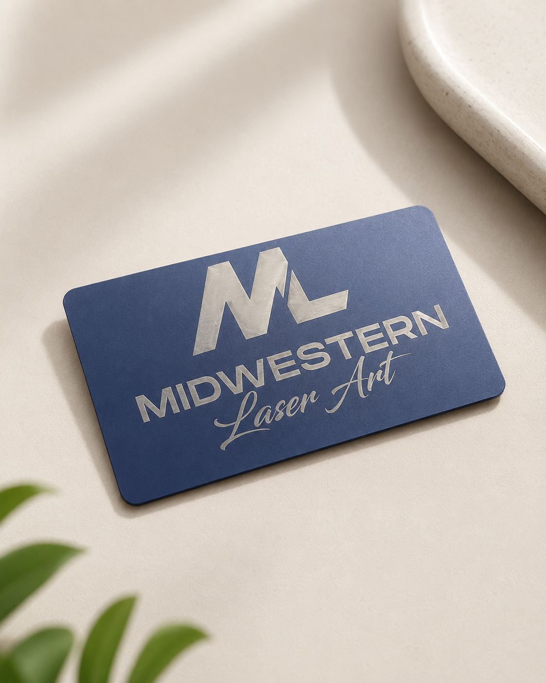 A rectangular blue card with silver foil branding reading 'MIDWESTERN Laser Art' and a stylized 'M' logo, photographed on a warm beige surface with soft shadows and partial green leaves in the corner.