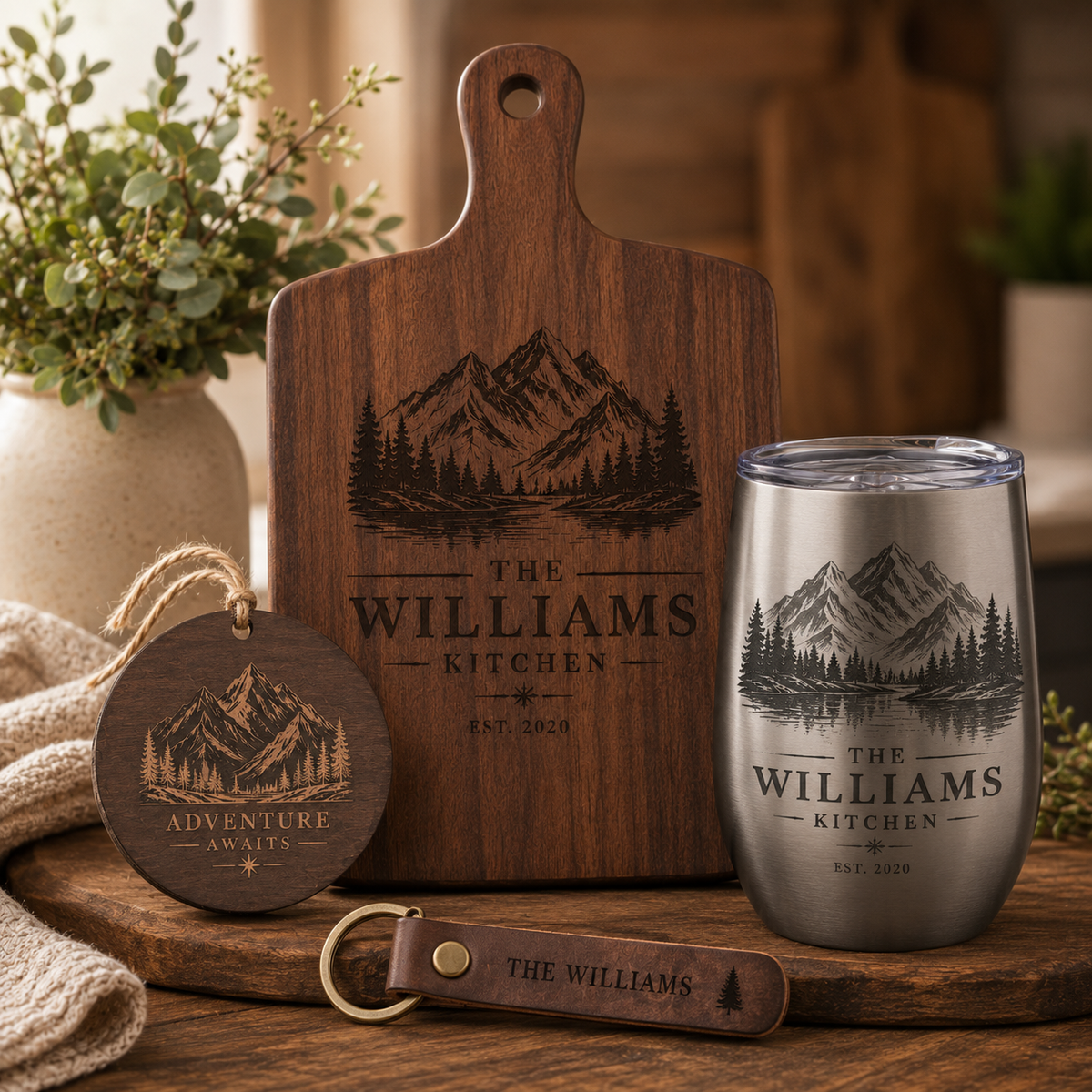 Personalized wooden and stainless steel kitchen-themed gift set on a rustic wooden table, including a cutting board, tumbler, round keychain, and leather key fob, all engraved with mountain artwork and the text 'The Williams Kitchen' and 'Adventure Awaits'.