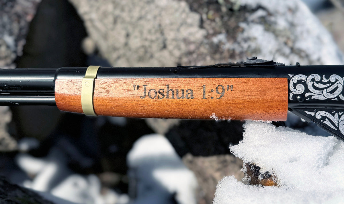 Wooden handle of a rifle with 'Joshua 1:9' engraving on a snowy background