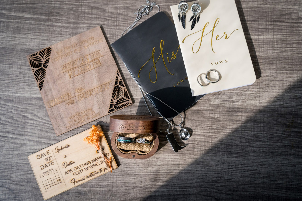 A styled flat-lay of wedding stationery and accessories on a gray wood background, including a kraft paper invitation, a black card with gold script reading 'His', a white card with gold script reading 'Her Vows', rings, earrings, a bracelet, and a save-the-date card.