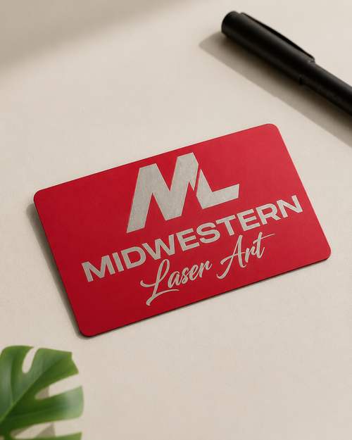 A red rectangular card with silver foil branding reading 'ML' and 'MIDWESTERN Laser Art' lies on a light beige surface, with a black pen in the top-right corner and a green leaf in the bottom-left corner.